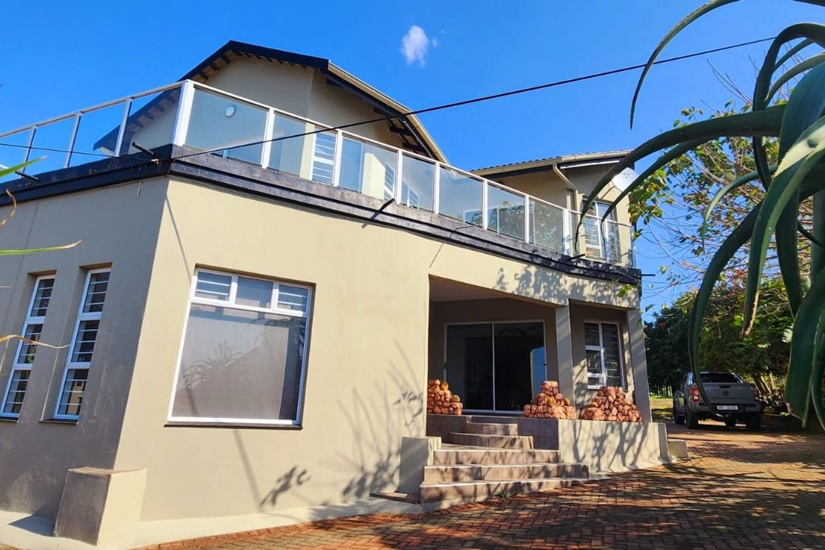 Rear view of a multi-level house featuring a spacious upper balcony with a glass balustrade, paved driveway, and tiled entrance steps.