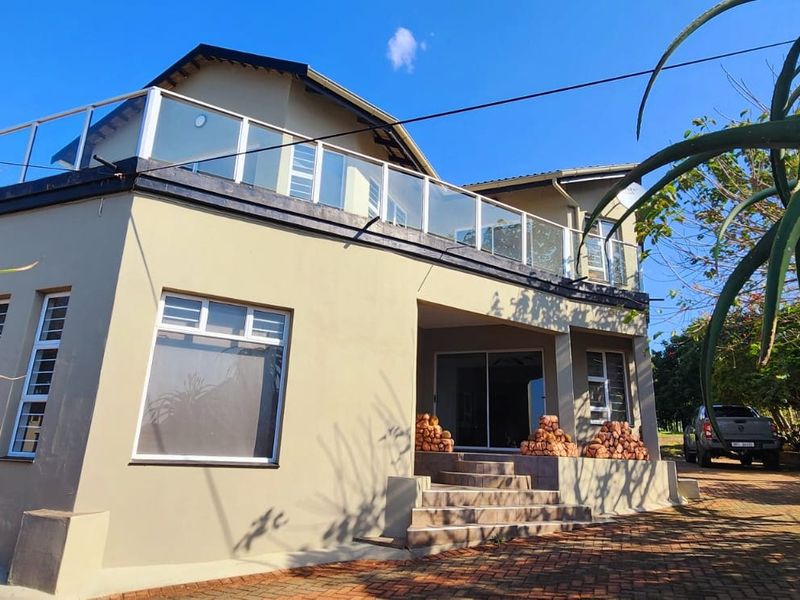 Rear view of a multi-level house featuring a spacious upper balcony with a glass balustrade, paved driveway, and tiled entrance steps.