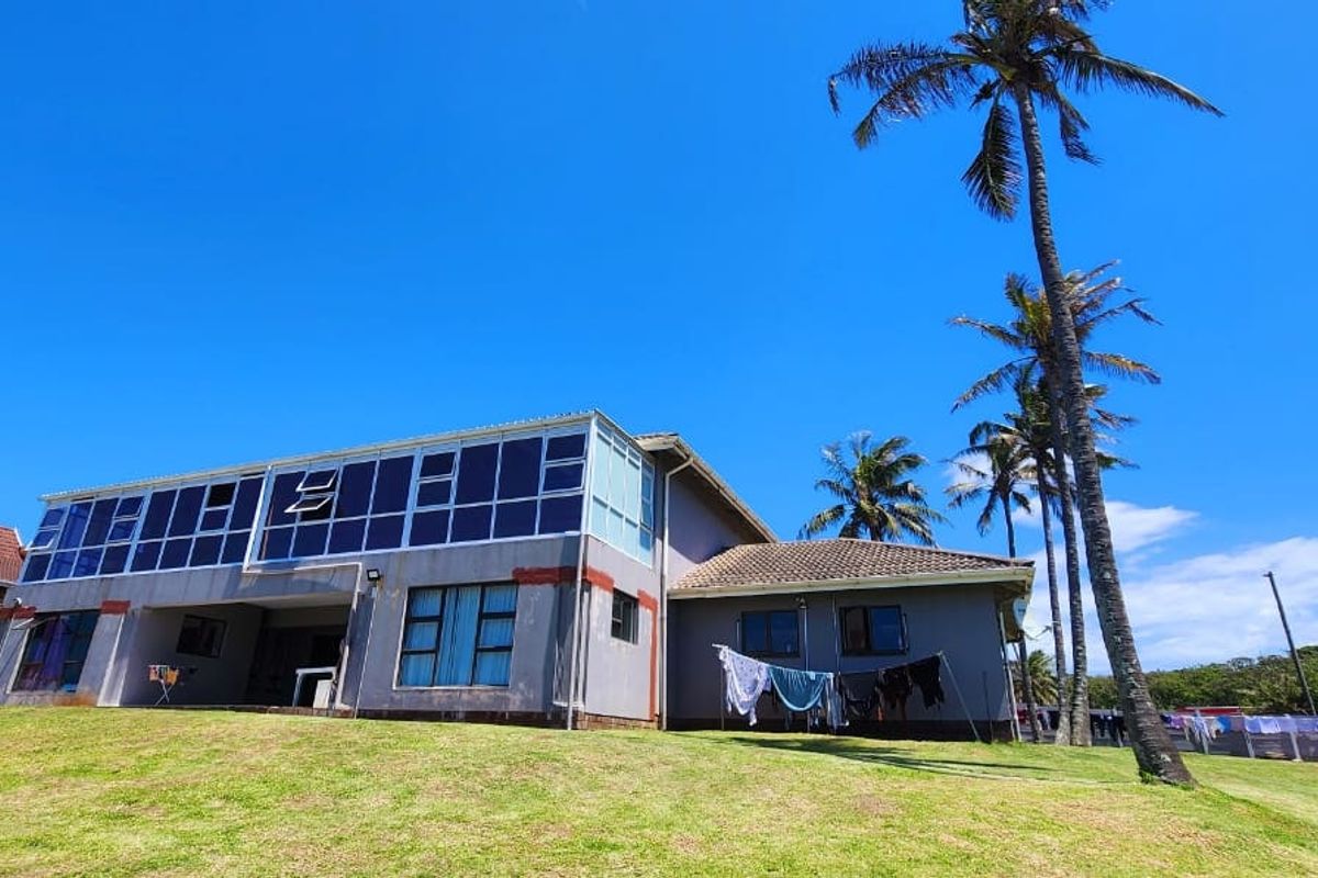 A rear view of the house featuring a large upper-level glass-enclosed balcony, tiled roof, and expansive lawn area with mature palm trees.