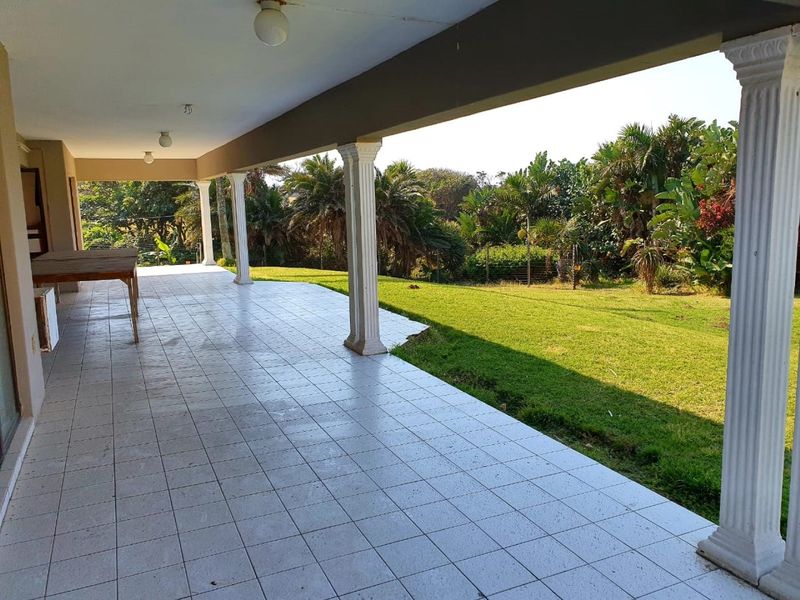 A spacious covered patio featuring tiled flooring and decorative pillars, overlooking a large, well-maintained garden area.