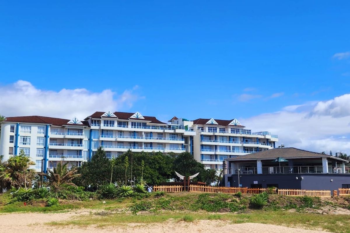 A wide exterior view of a multi-story apartment complex featuring balconies and a separate clubhouse structure, situated near the beach.