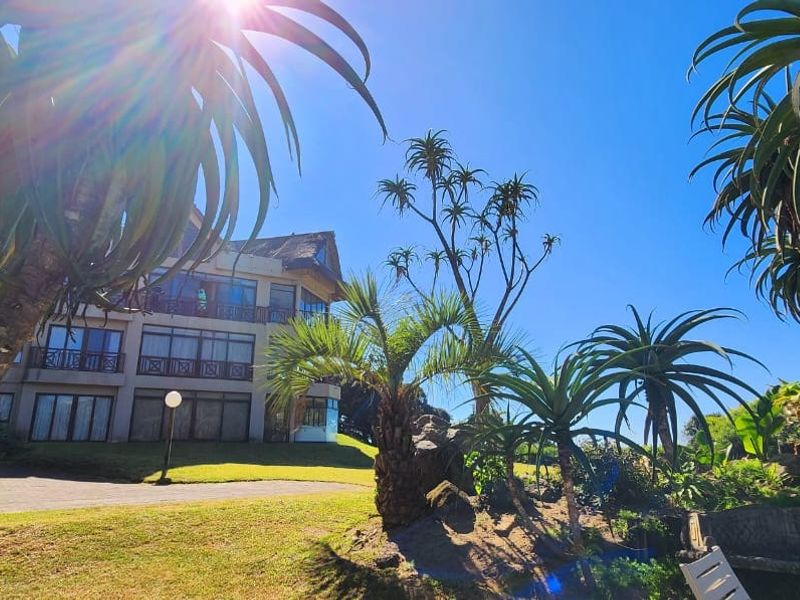 A view of the apartment building exterior featuring landscaped gardens, mature trees, and paved pathways under a clear blue sky.