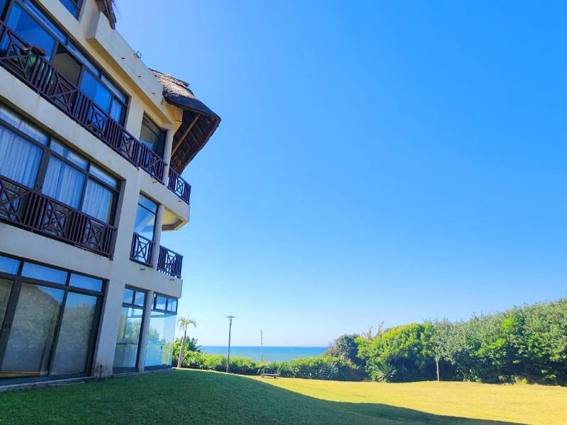 Exterior view of the apartment building facade featuring balconies and large glass sliding doors, overlooking a landscaped lawn and coastal scenery.
