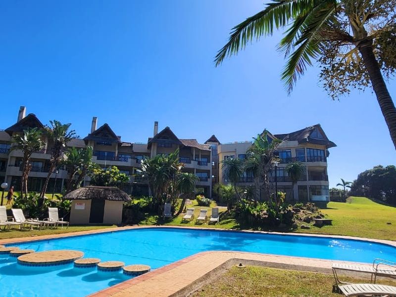 A well-maintained communal swimming pool area set within landscaped grounds, featuring a large pool and views of the apartment complex architecture.