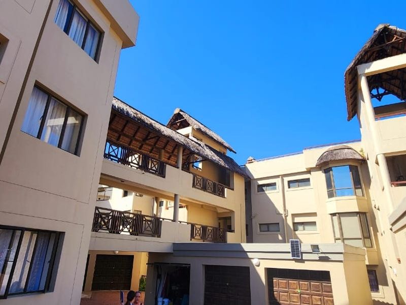 Exterior view of a multi-story apartment complex featuring thatched roof accents, balconies with wooden railings, and integrated garage doors.