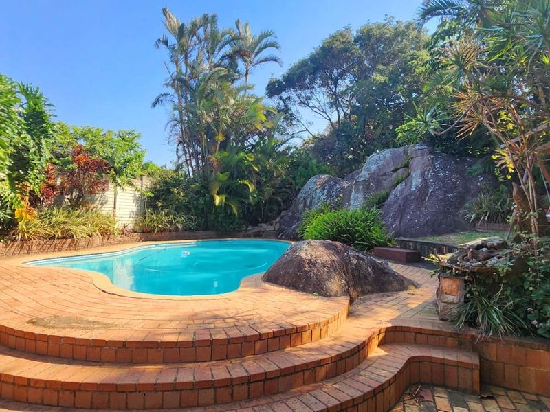 A landscaped outdoor pool area featuring a swimming pool, brick paved surround, and natural rock formations set within a lush garden environment.