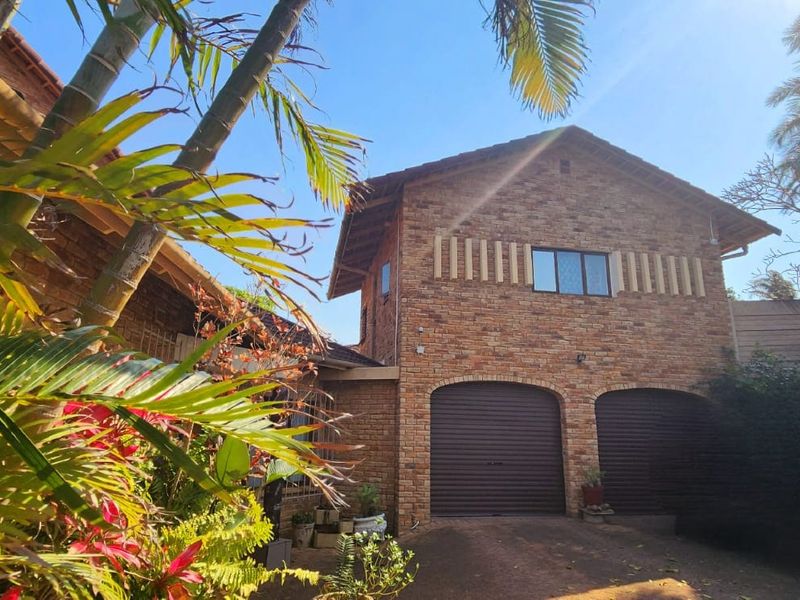 Front view of a double-story face brick house featuring a double garage with roll-up doors and established garden surroundings.