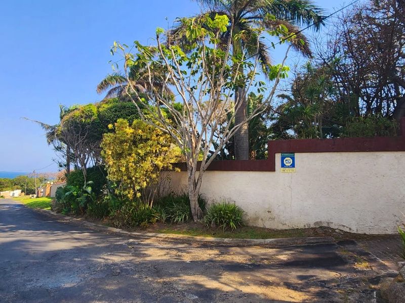 A street-level view of the property boundary wall, featuring established landscaping and security signage along the roadside.
