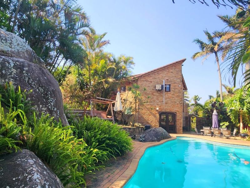 A private outdoor pool area featuring a brick-clad building, paved surrounds, and established garden landscaping with natural rock features.