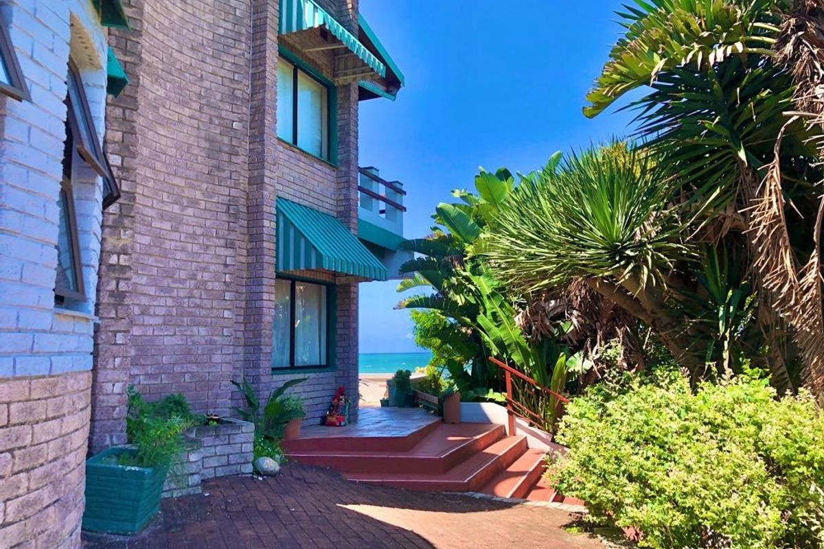 Rear exterior view of a multi-story brick house featuring a paved patio, staircase, and awnings, with a glimpse of the ocean in the background.