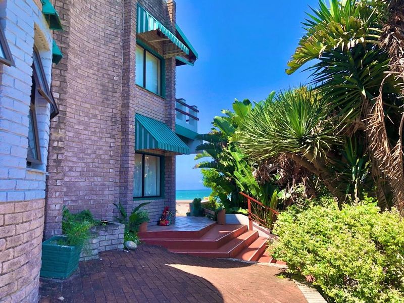 Rear exterior view of a multi-story brick house featuring a paved patio, staircase, and awnings, with a glimpse of the ocean in the background.