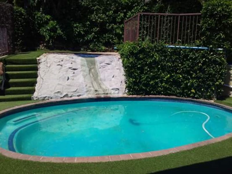 Outdoor pool area featuring an oval swimming pool, artificial grass landscaping, a decorative rock water feature, and perimeter metal fencing.