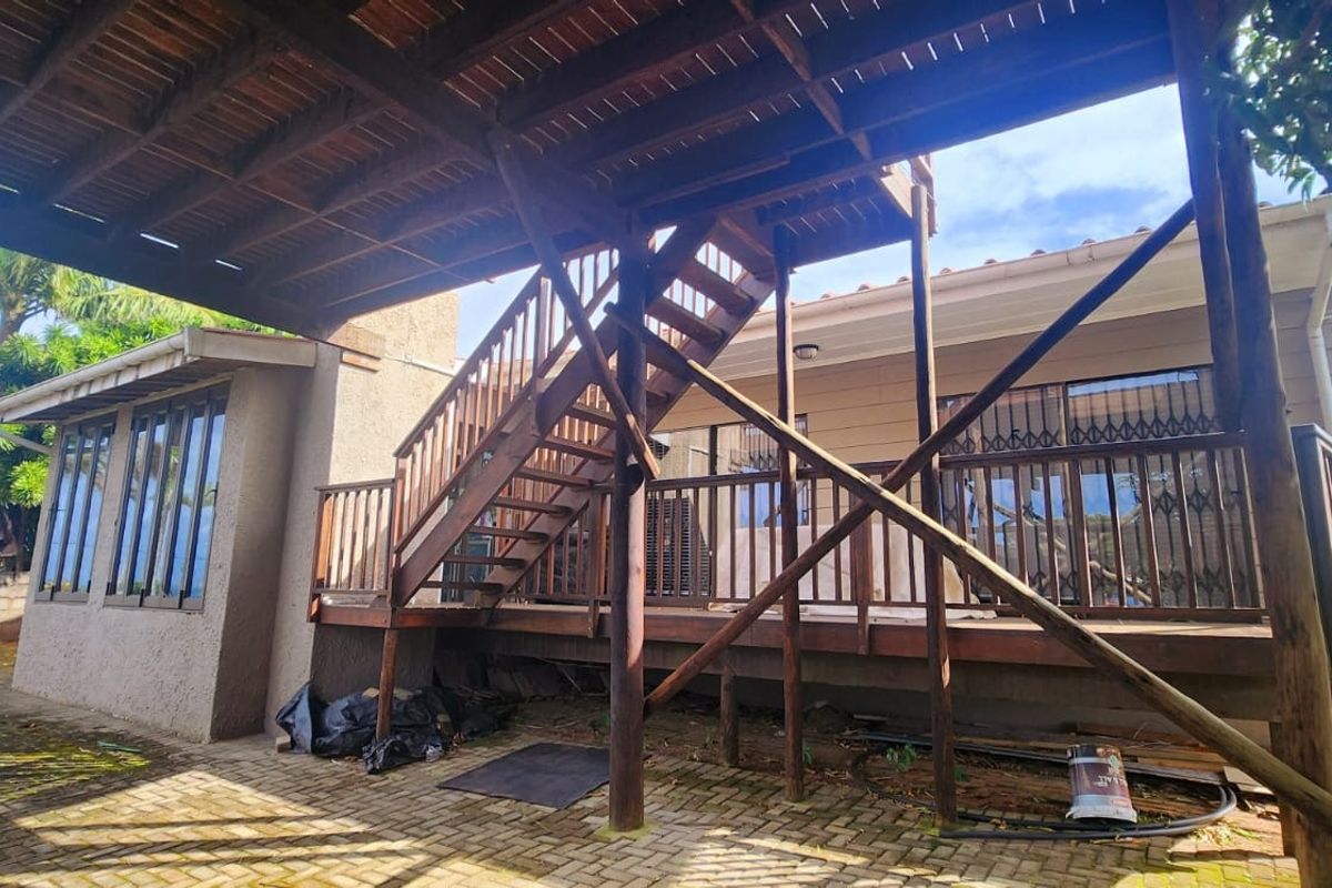 Rear exterior view featuring a raised timber deck with an external staircase, paved patio area, and security gates on the building access points.