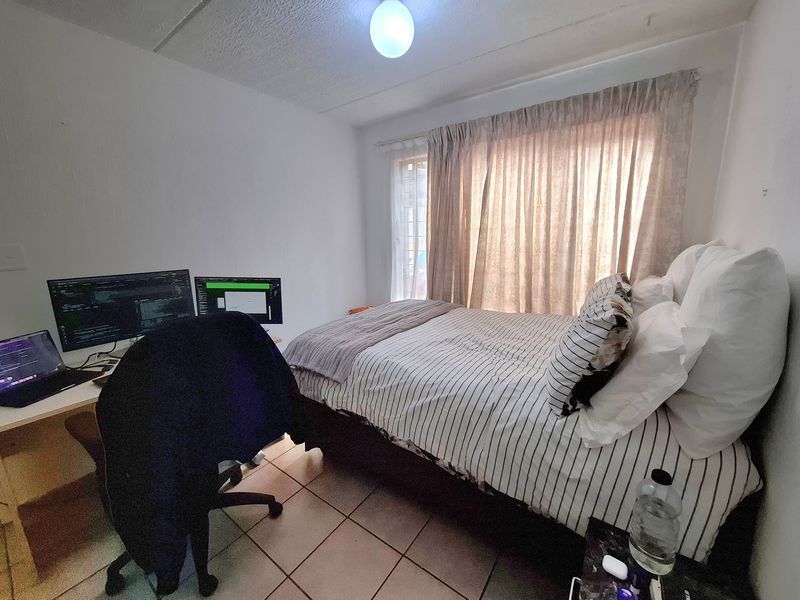 A compact bedroom featuring tiled flooring and a window with curtain tracks, currently utilized as a combined sleeping and workspace area.