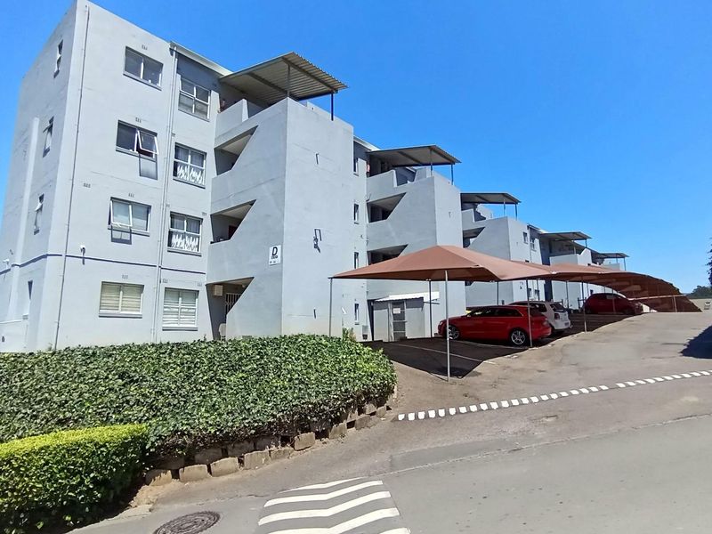 Exterior view of a multi-story apartment complex featuring covered parking bays, paved driveway, and established garden hedging.