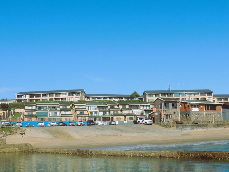 A view of a multi-story apartment complex situated directly on the beachfront, featuring a concrete boat ramp and stone masonry architectural details.