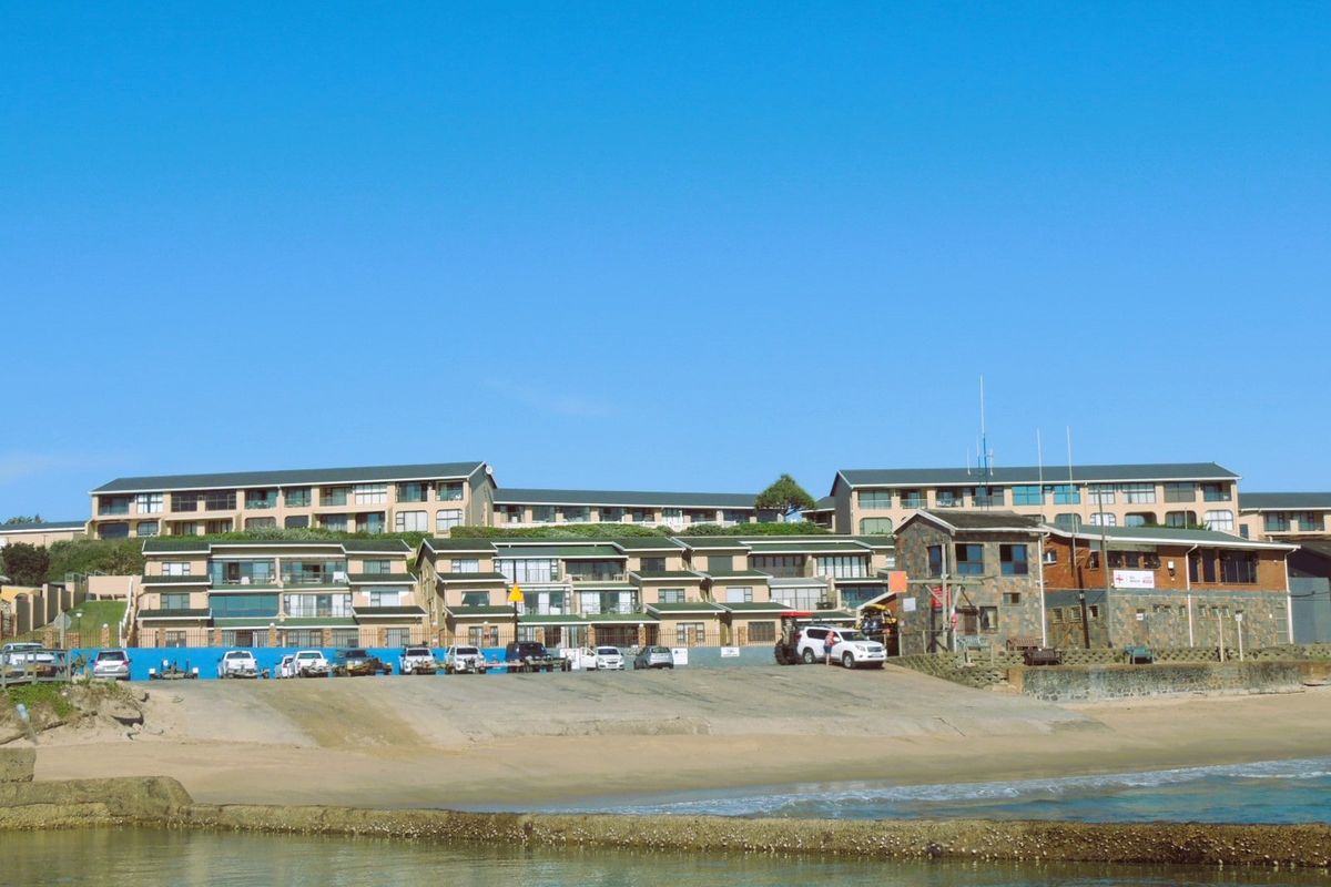 A view of a multi-story apartment complex situated directly on the beachfront, featuring a concrete boat ramp and stone masonry architectural details.