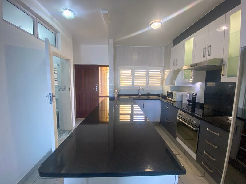 Modern kitchen featuring a long granite breakfast bar, sleek two-tone cabinetry, built-in oven, extractor fan, and tiled flooring with ample natural light.