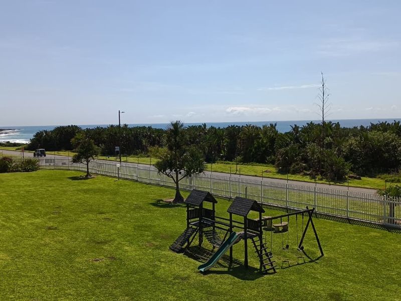 Elevated view overlooking a landscaped communal garden, playground area, and coastal road with ocean views in the distance.