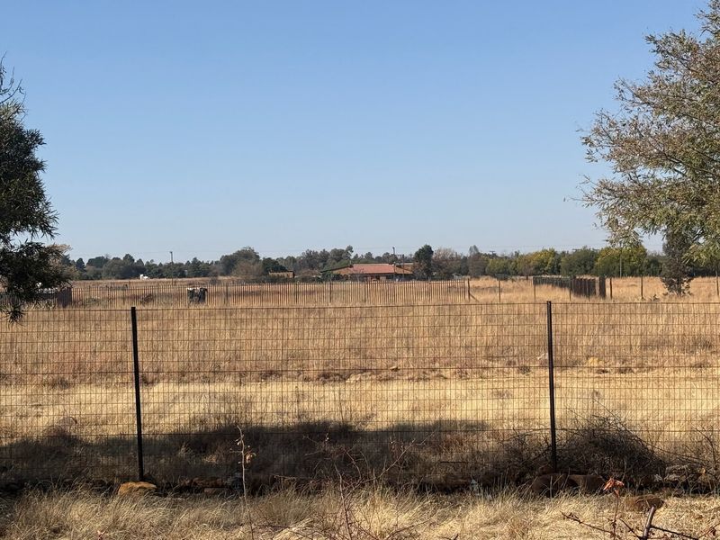 A wide view of an expansive, dry grassy field enclosed by wire fencing, with distant trees and structures visible on the horizon.