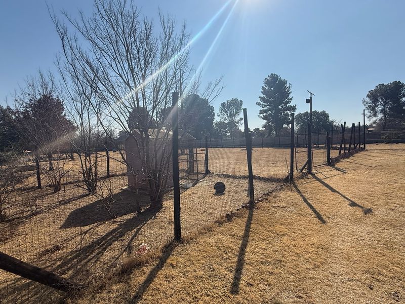 A large, fenced outdoor area featuring dry grass, mature trees, and a small shed structure, suitable for various outdoor uses.