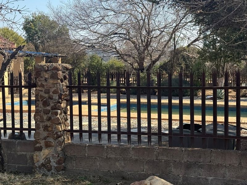 View of a swimming pool area enclosed by a metal security fence with stone-clad pillars and surrounding garden landscape.