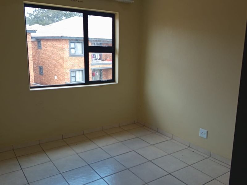 An empty bedroom featuring neutral wall paint, tiled flooring, and a large window providing natural light and a view of the surrounding complex.