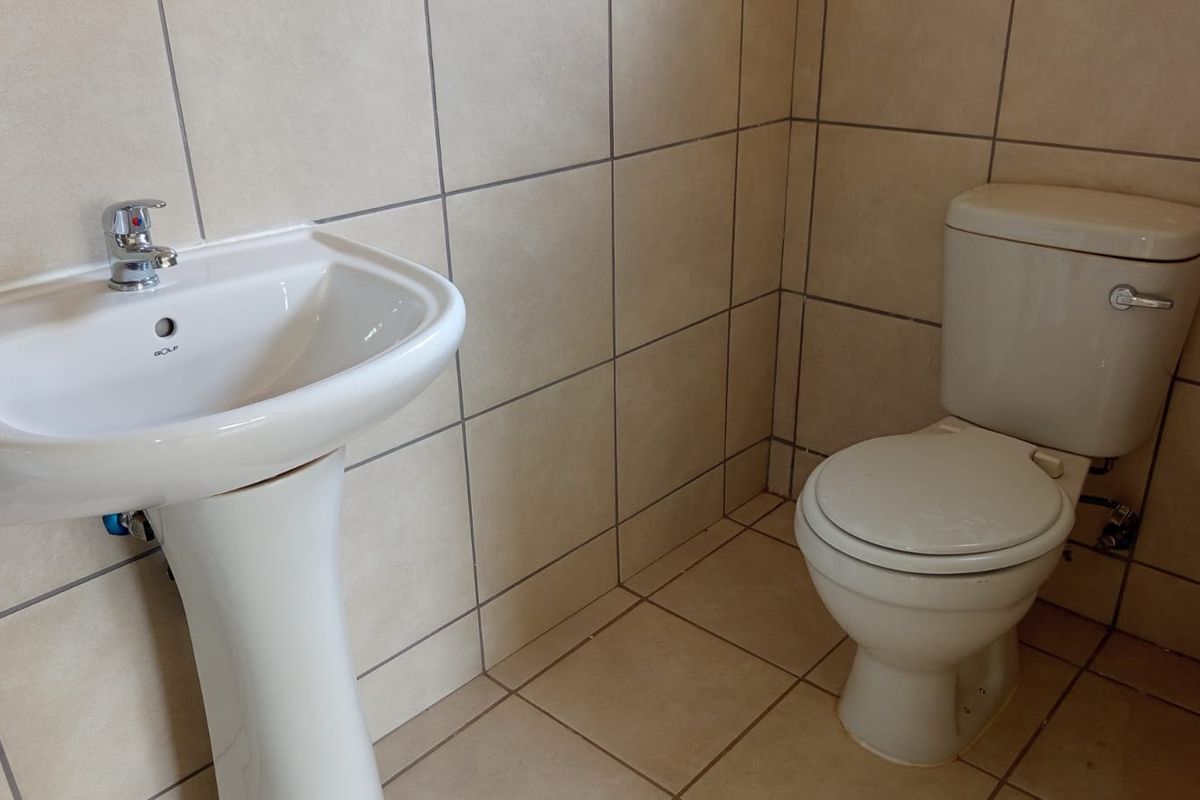 A functional bathroom featuring a pedestal washbasin with chrome faucet and a standard ceramic toilet, finished with neutral wall and floor tiling.