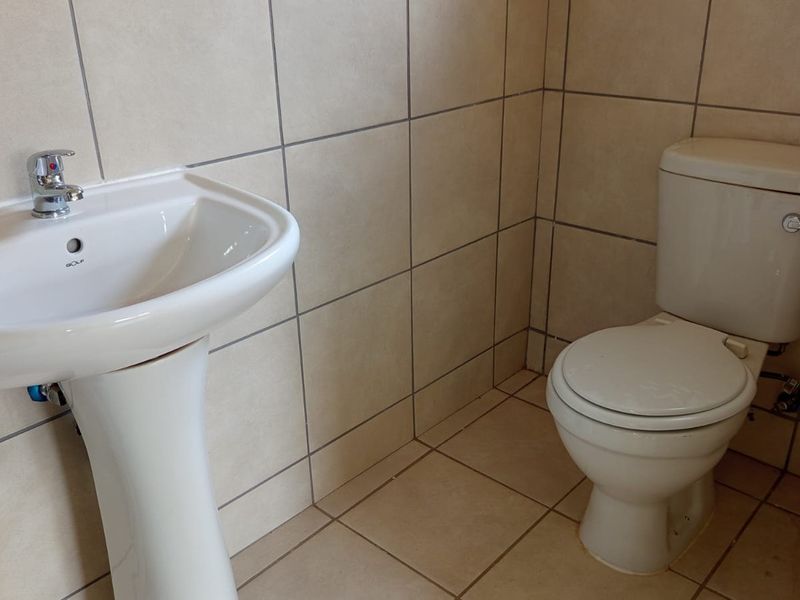 A functional bathroom featuring a pedestal washbasin with chrome faucet and a standard ceramic toilet, finished with neutral wall and floor tiling.