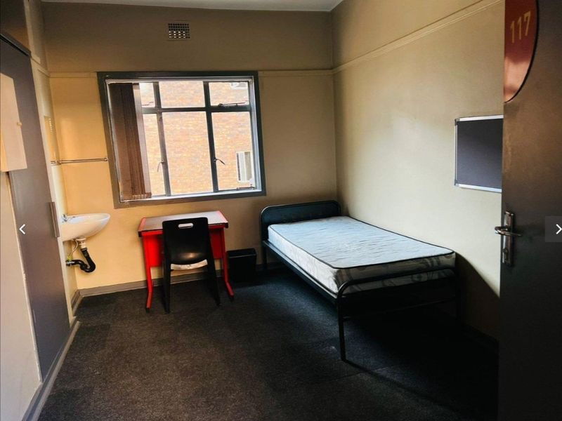 A compact bedroom featuring a wall-mounted basin, metal-framed window, and built-in storage, offering a functional space with potential for cosmetic updates.