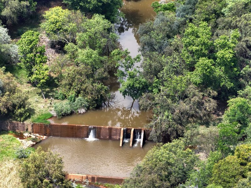 An aerial view of a natural river flowing through the property, featuring a concrete weir structure surrounded by mature trees and vegetation.