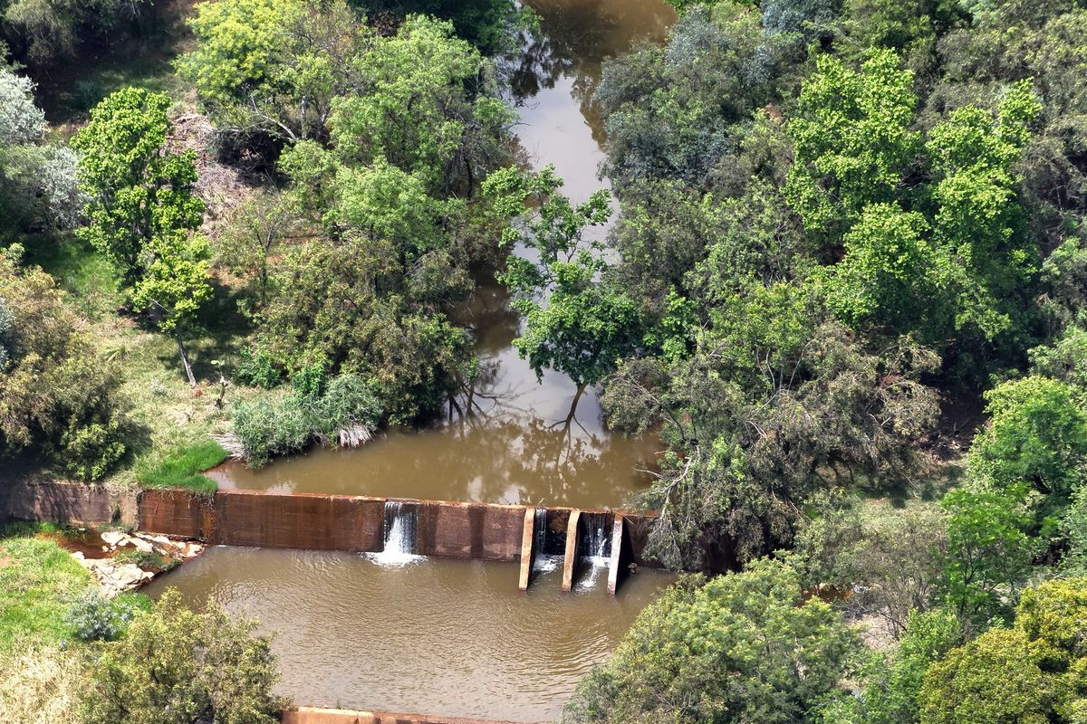 An aerial view of a natural river flowing through the property, featuring a concrete weir structure surrounded by mature trees and vegetation.