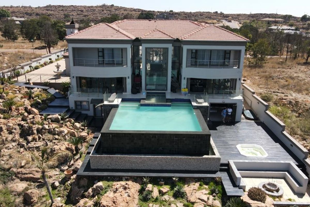 Aerial view of a modern luxury residence featuring a prominent infinity-edge swimming pool, expansive decking, built-in fire pit, and a jacuzzi set in a rocky landscape.