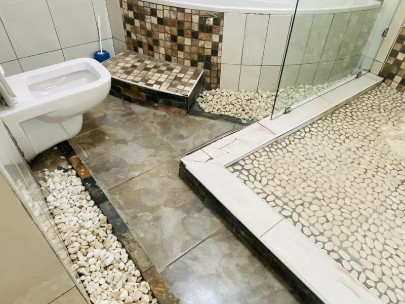 Bathroom featuring a corner bathtub, glass shower enclosure, and wall-mounted toilet, with decorative pebble and mosaic tile detailing requiring maintenance.