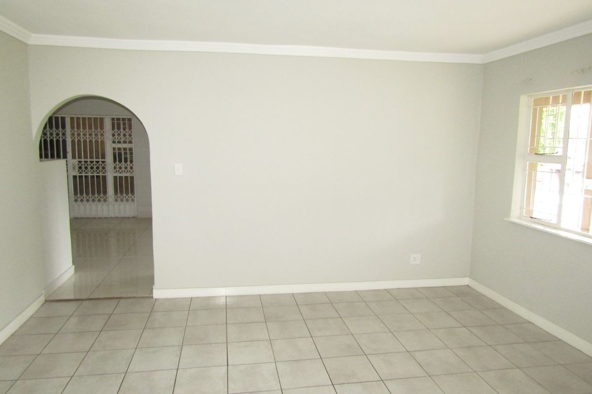 A bright, empty living room featuring neutral tiled flooring, coving, and an arched doorway leading to an adjacent space with security gates.