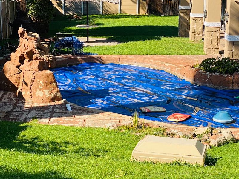 A backyard pool area featuring a rock-accented swimming pool, paved surround, and a covered patio area adjacent to a landscaped garden.