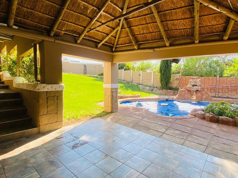 Covered outdoor patio featuring a thatched roof and stone pillars, overlooking a swimming pool with a rock water feature and landscaped garden.