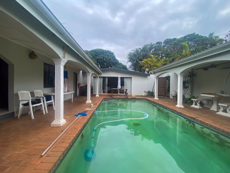 Outdoor pool area featuring a covered patio with decorative columns and brick paving, surrounded by established garden greenery.