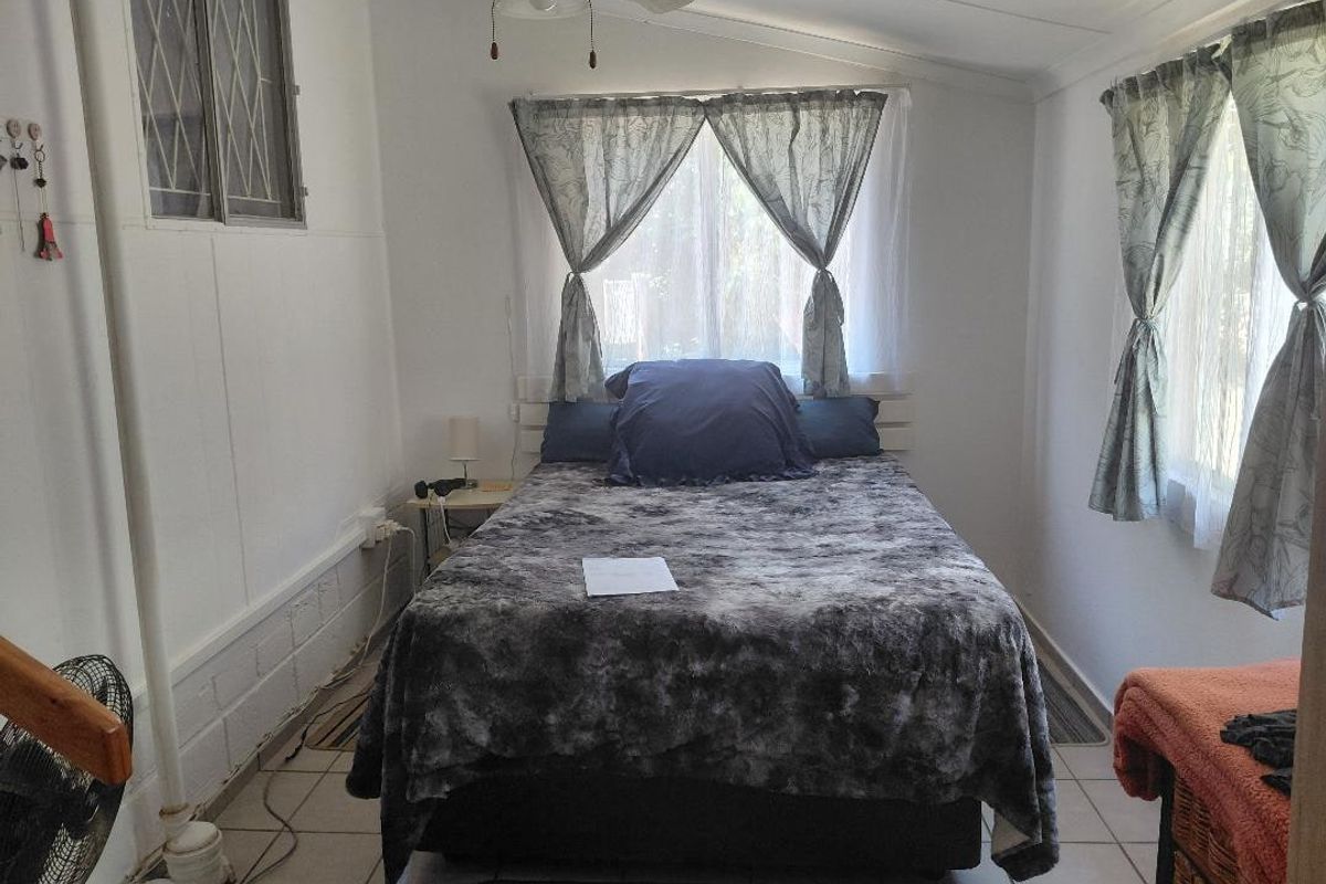A bedroom featuring tiled flooring, a ceiling fan, and security bars on the window, with an exposed plumbing pipe along the wall.