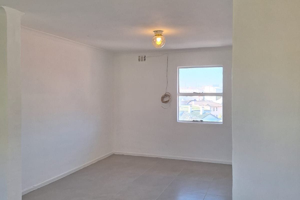 A bright, empty living area featuring neutral floor tiles, a single ceiling light fixture, and a window with a view of the surrounding neighborhood.