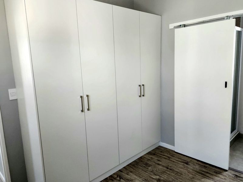 A clean bedroom interior featuring modern white built-in wardrobes, wood-look laminate flooring, and a sliding door leading to an adjacent space.