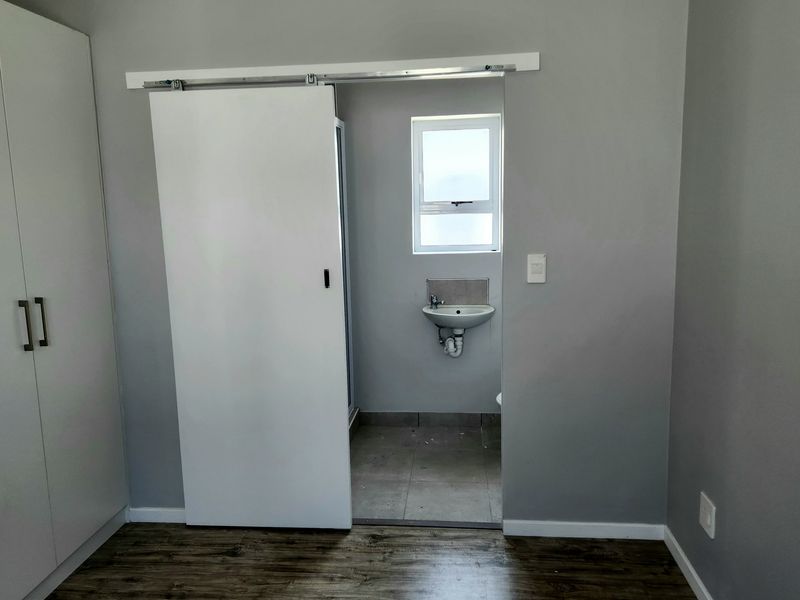 A clean bedroom featuring modern laminate flooring, a built-in wardrobe, and a sliding door leading to an en-suite bathroom with a wall-mounted basin.