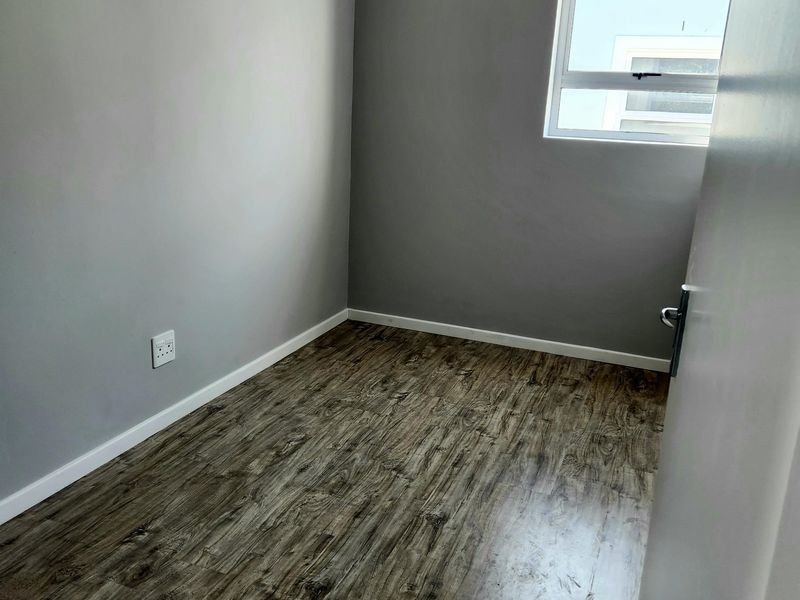 A clean, empty bedroom featuring modern grey walls, wood-look laminate flooring, and a single casement window providing natural light.