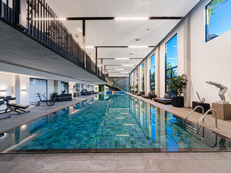 A modern, high-end indoor swimming pool area featuring a long lap pool, mezzanine level, and expansive windows providing natural light.