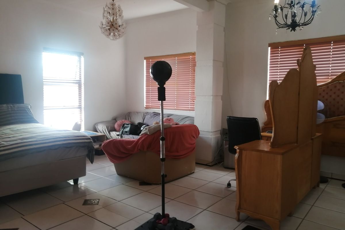 A spacious bedroom featuring tiled flooring, dual chandeliers, and large windows with fitted blinds, offering a versatile layout for living and sleeping.