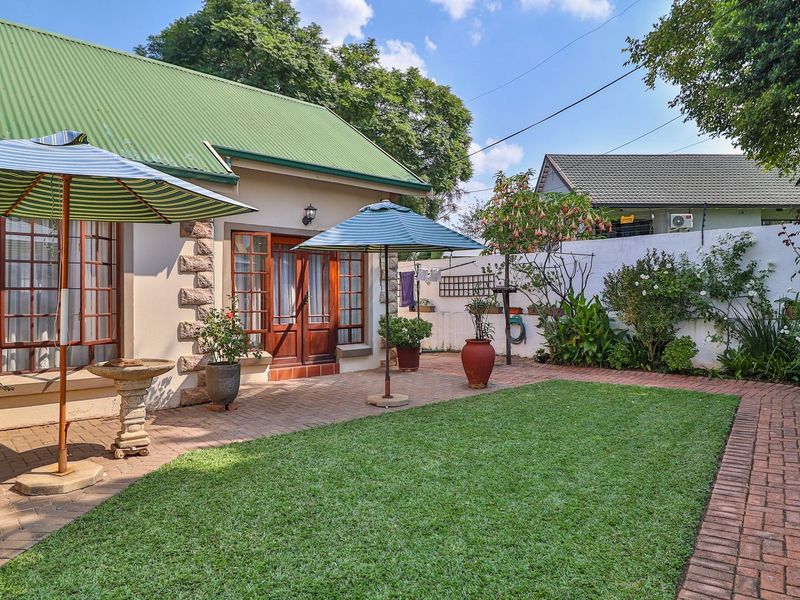 A well-maintained rear garden featuring a brick-paved patio, a neat lawn, and a charming house exterior with stone accents and a green metal roof.