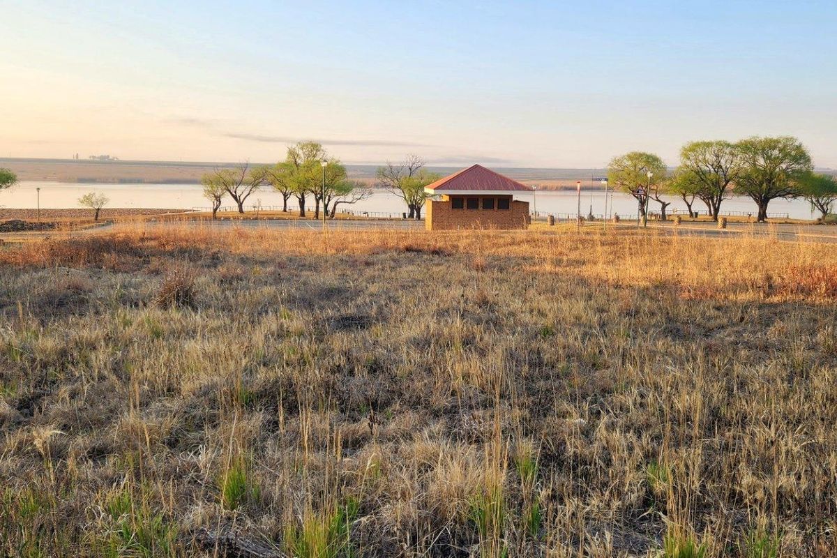 A scenic view of a vacant residential plot overlooking a tranquil lake with mature trees and a small structure in the distance.