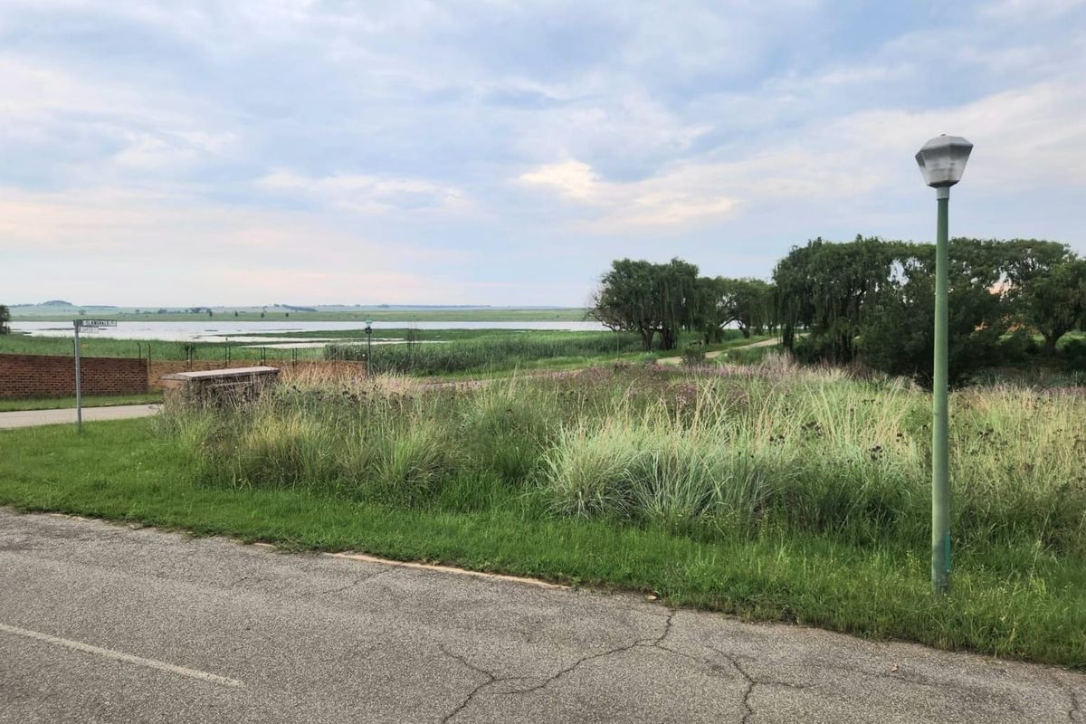 A view of a vacant residential plot featuring natural landscape, a brick boundary wall, and a street lamp overlooking a nearby water body.