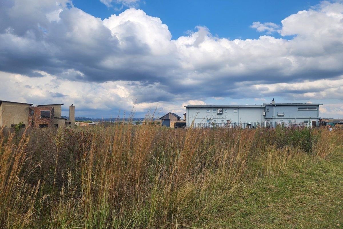 A view of a vacant residential plot featuring tall grass with surrounding residential properties in the background.