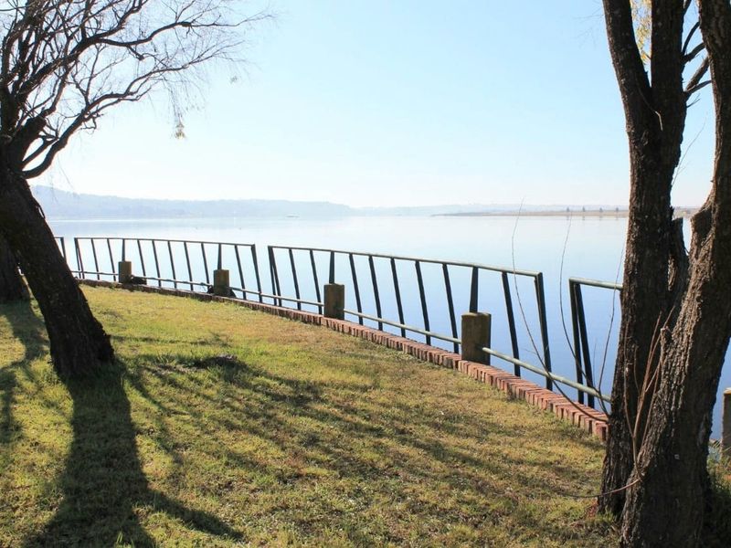 A scenic waterfront view featuring a grassy bank, a brick retaining wall, and a damaged metal safety railing overlooking the water.